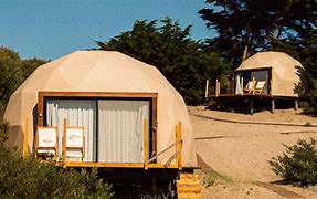 Two glamping structures at a sandy beach with a deck and chairs.