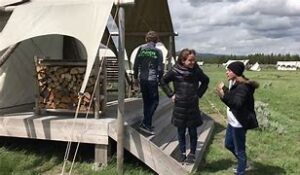 Under Canvas Yellowstone – Wyoming, USA 2 People on stepsd talking to each other at a glamping tent structure with wood stacked on a platform above the ground.