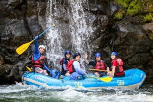 Ultimate Outdoor & Adventure Guide 1 People canoeing in a raft wearing life vests on a river near a waterfall and steep cliff