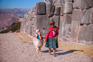 Woman walking a llama along a stone wall at an ancient ruin in Peru’s Sacred Valley, with mountains in the background.