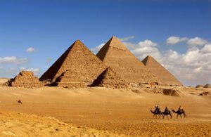 Huge pyramids in a desert with people riding on camels with a blue sky.