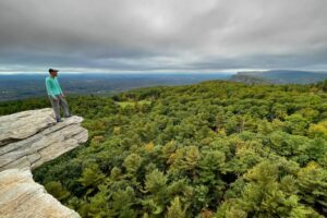 Mohonk Preserve, NY: Hiking, Climbing & Hudson Valley Beauty 2 Person standing on a cliff edge overlooking a lush valley filled with dense forest.