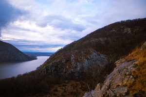 Scenic mountain overlook above the Hudson River featuring steep rocky cliffs, forested hillsides, rugged hiking terrain and a winding river beneath a dramatic cloudy sky in New York's Hudson Highlands.
