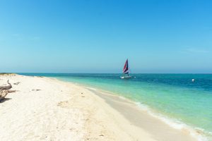 Beautiful white-sand beach with crystal-clear turquoise waters, gentle waves, a colorful sailboat offshore, and a bright blue sky creating a peaceful tropical coastal scene.
