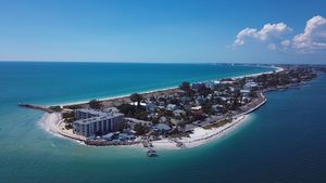 Aerial view of a Florida barrier island featuring white-sand beaches, turquoise Gulf waters, waterfront homes, coastal roads, and a long shoreline stretching into the distance.