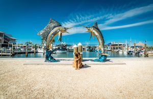 Two large sailfish sculptures overlooking a sunny waterfront marina with docked boats, a dog sitting between the sculptures, and a bright blue Florida sky.