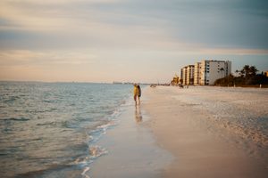 Caladesi Island State Park: A Hidden Paradise Near Clearwater Beach 2 Peaceful Florida Gulf Coast beach at sunset, featuring gentle waves, soft white sand, two people standing near the shoreline, and beachfront buildings surrounded by palm trees.