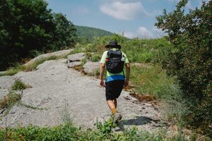 Harriman State Park, NY: Lakes, Trails & Wilderness Just Outside NYC ✨ 2 Hiker with a backpack walking across a rocky trail surrounded by green vegetation and forested hills at Harriman State Park, New York.
