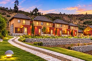 Scenic mountain lodge retreat featuring rustic-style cottages with stone accents, warmly illuminated windows, private balconies, landscaped gardens, and a winding stone pathway. Beautiful mountain hillsides and a colorful sunset sky create a peaceful atmosphere inspired by Colca Canyon, Peru.