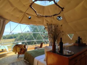 Inside a cozy glamping dome with a bed, counter, and two chairs facing a large window that opens to a deck.