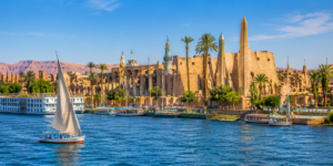 Luxor Temple – Heart Of Ancient Thebes 13 Daytime view of the Nile River at Luxor, Egypt, with a traditional felucca sailing past palm trees and the ancient Luxor Temple complex.