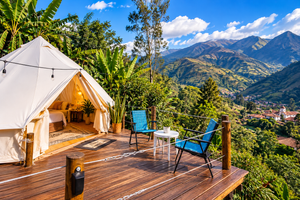 Highland Glamping in Antioquía, Peru: Mountain Retreats & Scenic Adventures 1 Luxury white canvas glamping tent on a spacious wooden deck overlooking lush green Andean mountains and a picturesque village, featuring comfortable outdoor chairs, tropical vegetation, and beautiful blue skies, illustrating highland glamping near Antioquía, Peru.