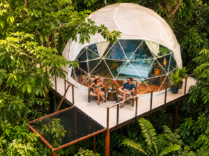 Couple relaxing on the deck of an elevated geodesic dome surrounded by lush tropical rainforest.
