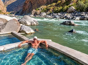 Man wearing sunglasses and a white cap relaxing in a clear turquoise pool beside a flowing river, surrounded by large boulders, rugged canyon cliffs, and beautiful Andean mountain scenery near Llahuar, Colca Canyon, Peru.