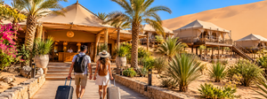 Luxury Tents in Huacachina Oasis: Desert Glamping & Adventures in Peru 6 Two travelers carrying luggage walk toward a luxury desert glamping resort featuring safari-style tents, palm trees, wooden walkways, and golden sand dunes near Huacachina Oasis, Peru.