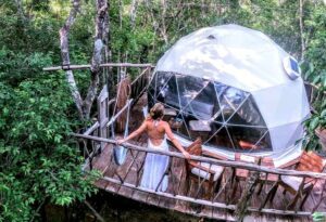 White glamping dome with a woman standing on the deck, surrounded by chairs and a hammock, featuring a large window.
