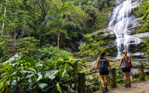 Two women hikers enjoying a tropical rainforest waterfall surrounded by lush vegetation.