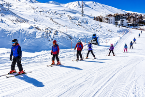 La Parva Mountain Retreat: A High-Altitude Escape in the Andes 2 Group of skiers wearing colorful winter jackets, helmets, and ski equipment descending a wide, snow-covered mountain slope, with ski lifts, alpine resort buildings, and spectacular snowy mountain peaks in the background.