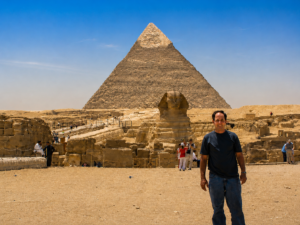 Traveler standing near the Great Pyramid and Sphinx at Giza, Egypt under a bright blue sky with ancient ruins and desert scenery in the background.