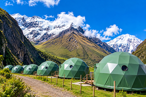 Green geodesic glamping domes lined along a mountain pathway, surrounded by lush vegetation, towering Andean mountains, and spectacular snow-covered peaks beneath a bright blue sky.