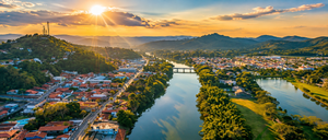 Guararema Riverside Glamping, Brazil: A Peaceful Escape by the Water 1 Wide panoramic aerial view inspired by Guararema, São Paulo, Brazil, featuring a winding river, charming town, lush green hills, bridges, and golden sunset rays illuminating the countryside.