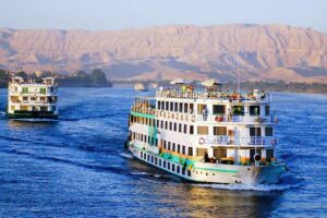 Two cruise shipswith people on the ships cruising on the Nile River with mountains in the background