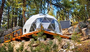 White geodesic glamping dome on a wooden platform surrounded by pine trees and large rocks in a secluded mountain forest.