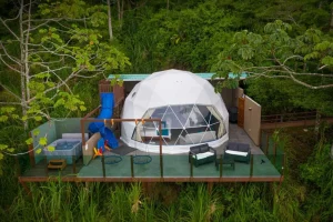 ✨ Rio De Janeiro Glamping Hub: Luxury Nature Stays in Brazil’s Most Vibrant City 20 White geodesic glamping dome on an elevated wooden platform surrounded by lush green forest, with outdoor chairs, a hot tub, and recreational amenities.