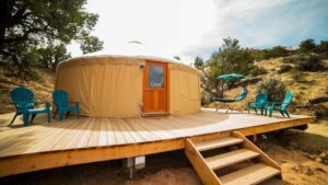 Tan glamping yurt on a raised platform surrounded by trees, chairs, and sandy ground.