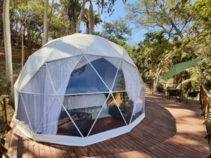 White glamping dome with windows and a wooden deck featuring chairs and a table surrounded by nature.