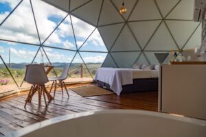 Interior of a glamping dome with a cozy bed, small table, two chairs, and a large window overlooking nature
