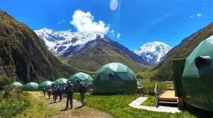 Green geodesic glamping domes nestled in a scenic Andean mountain valley near Cusco, Peru, with visitors walking along a grassy pathway, towering rocky mountains, and snow-capped peaks beneath a bright blue sky.
