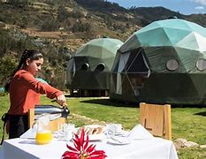Woman at a table and chairs with food and a couple glamping dfomes in a valley with mountains.