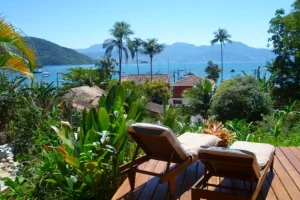 ✨ Rio De Janeiro Glamping Hub: Luxury Nature Stays in Brazil’s Most Vibrant City 21 Wooden sun loungers on a tropical garden terrace overlooking blue ocean waters, palm trees, coastal houses, and distant green mountains.