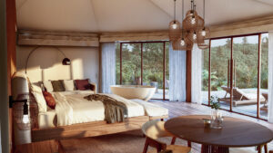 Interior of a glamping dome featuring a bed, table and chairs, and a hot tub with two large windows opening to a deck with lounge chairs and a rainforest view
