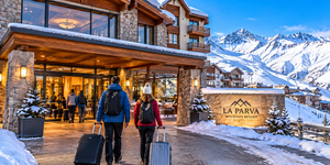 La Parva Mountain Retreat: A High-Altitude Escape in the Andes 9 Travelers wearing winter jackets and carrying luggage walk toward the warmly illuminated entrance of a mountain resort in La Parva, Chile, surrounded by snow-covered alpine buildings, towering Andean peaks, and beautiful winter scenery.