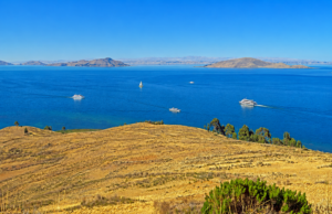 Scenic high-altitude lake surrounded by rolling golden hills and distant islands, featuring colorful boats crossing the deep blue waters beneath a clear sky in the Peruvian Andes.