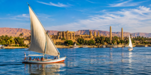 Luxor Temple – Heart Of Ancient Thebes 15 Traditional Egyptian felucca sailing on the Nile River near Luxor, with blue water, palm-lined shores, ancient temples and desert mountains in the background.