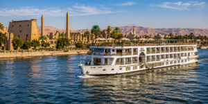 Luxor Temple – Heart Of Ancient Thebes 14 Nile River cruise ship sailing near Luxor, Egypt, with blue water, palm trees, ancient Egyptian temples and desert mountains in the background.