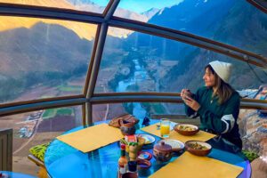 Woman enjoying a meal at a table inside a glamping dome, overlooking a scenic valley through large panoramic windows.