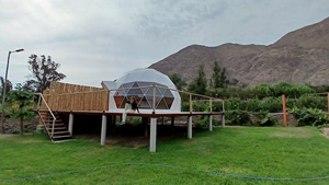 White geodesic glamping dome on an elevated wooden platform overlooking a green lawn, lush vegetation, and rugged mountains in Peru's Lunahuaná Valley, with a visitor relaxing on the deck.