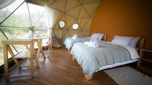 Interior of a luxury geodesic glamping dome featuring two comfortable beds, wooden flooring, a dining table, large panoramic windows, and scenic natural views.
