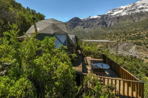 Dome-style glamping accommodation with a wooden deck surrounded by greenery and dramatic snow-capped Andes Mountains in Cajón del Maipo, Chile.