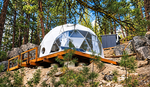 Glamping In Cajón Del Maipo: Andes Luxury Camping Near Santiago, Chile 4 White geodesic glamping dome with panoramic windows on an elevated wooden deck surrounded by pine trees, natural rocks, and peaceful forest scenery.