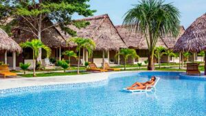Woman lying in a pool with huts and lounge chairs with trees.