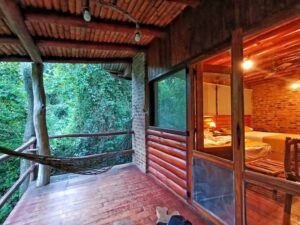 🌅 Riverside Glamping in Puerto Iguazú: Jungle Comfort by the Water 3 Wooden building in a rainforest with a covered porch featuring a hammock surrounded by lush green vegetation.