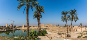 Karnak Temple – Luxor, Egypt: Gateway To Ancient Wonders 1 Panoramic view of Karnak Temple in Luxor, Egypt, featuring the Sacred Lake, palm trees, ancient ruins, temple structures, and obelisk beneath a clear blue sky.