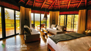 “Interior of a glamping structure featuring a bed, love seat, large windows, and a deck outside with lounge chairs overlooking lush rainforest