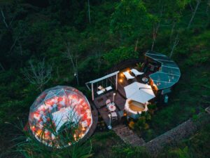 Glamping dome on a platform with a spa, swing set, and lounge chairs in a forest with vegetation.