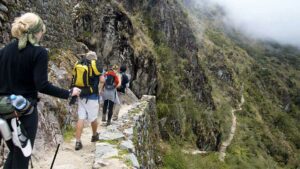 several people with backpacks walking up a trail with steep mountains and a rock wall.